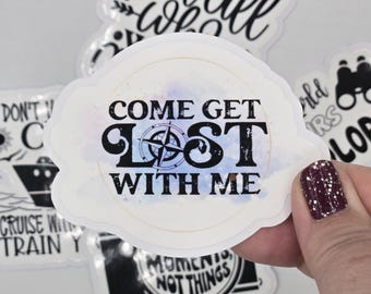 Könnte beinhalten: Weißer Aufkleber mit dem Text "COME GET LOST WITH ME" in Schwarz, mit einem Kompassrosen-Design. Weitere Aufkleber mit Reisemotiven sind im Hintergrund zu sehen.