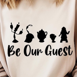 Puede incluir: Una sudadera beige con una silueta negra de personajes de la película de Disney La Bella y la Bestia. El texto "Be Our Guest" está impreso debajo de las siluetas.