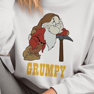 Puede incluir: Sudadera blanca con una imagen de dibujos animados de Gruñón de Blancanieves y los Siete Enanitos. Gruñón se apoya en un pico y el texto "GRUMPY" está impreso debajo de la imagen.