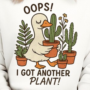 Puede incluir: Sudadera blanca con un pato de dibujos animados que lleva plantas en macetas. El texto "OOPS! I GOT ANOTHER PLANT!" está impreso en la parte delantera. El diseño presenta cactus verdes y otras plantas en macetas de terracota.