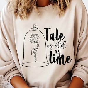 Puede incluir: Una sudadera beige con un gráfico negro de una rosa en una cúpula de vidrio y el texto "Tale as old as time".