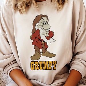 May include: A beige sweatshirt with a cartoon image of Grumpy from Snow White and the Seven Dwarfs. The image shows Grumpy with his arms crossed and a grumpy expression. The text "GRUMPY" is printed in yellow below the image.