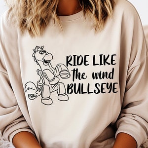 Puede incluir: Una sudadera beige con un contorno negro de un caballo y el texto "Ride like the wind Bullseye".