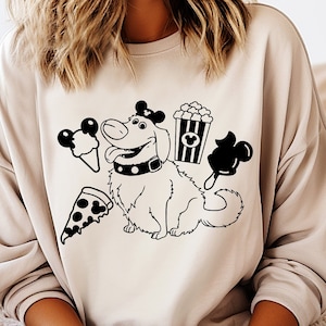 May include: A light beige sweatshirt with a black and white graphic print of a dog wearing a bow and holding a popcorn bucket, ice cream cone, and Mickey Mouse ears. The dog is surrounded by other Mickey Mouse themed food items.