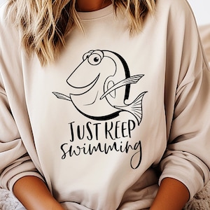 Può includere: Una felpa beige con un'illustrazione in bianco e nero di un pesce e il testo "Just Keep Swimming".