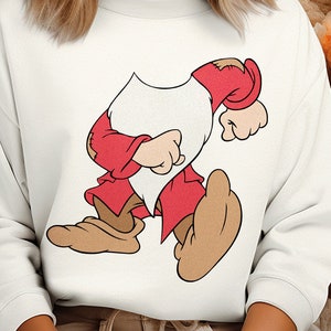 Pode incluir: Camisola branca com uma imagem de desenho animado de um anão a usar um chapéu vermelho e calças vermelhas. O anão está a andar e tem uma barba branca.