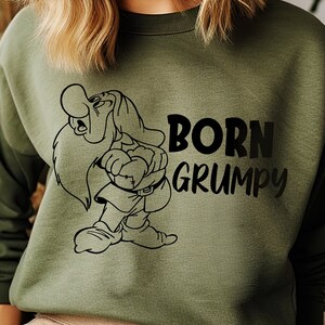 Puede incluir: Una sudadera verde con un gráfico negro de Grumpy de Blancanieves y los Siete Enanitos. El texto "Born Grumpy" está impreso debajo del gráfico.