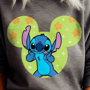 Pode incluir: Uma camisa de moletom cinza com um personagem Stitch azul usando um chapéu de orelha de Mickey Mouse. O chapéu é verde com flores laranja e brilhos brancos.