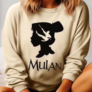 Puede incluir: Una sudadera beige con una silueta negra de una cabeza de mujer con un pájaro volando fuera de ella. El texto "MULAN" está impreso debajo de la silueta.