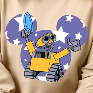 Puede incluir: Una sudadera beige con un personaje de robot de dibujos animados que lleva una visera azul y sostiene un disco azul. El robot está delante de una silueta de oreja de Mickey Mouse morada con estrellas blancas.
