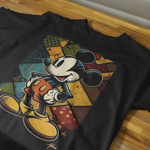May include: Black t-shirt with a colorful graphic of Mickey Mouse in a retro style. The graphic features Mickey Mouse standing with his arms crossed and a colorful geometric pattern behind him.