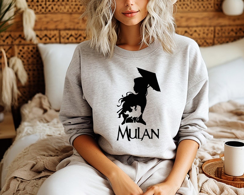 Op de afbeelding: Een grijs sweatshirt met een zwart silhouet van Mulan die een paraplu vasthoudt en het woord "MULAN" op de voorkant.