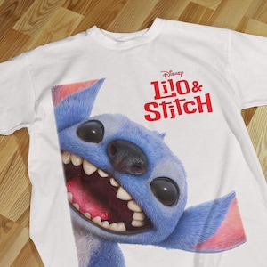 Può includere: T-shirt bianca con una grande grafica del personaggio Disney Stitch. Stitch è blu con grandi occhi neri, bocca rosa e grandi orecchie. Le parole "Disney Lilo & Stitch" sono stampate in rosso.
