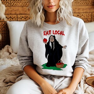 Puede incluir: Una sudadera gris claro con las palabras "EAT LOCAL" en rojo. El gr&aacute;fico presenta una bruja de dibujos animados sosteniendo una manzana roja y una cesta de manzanas. La bruja lleva una capa negra y est&aacute; de pie sobre hierba verde.