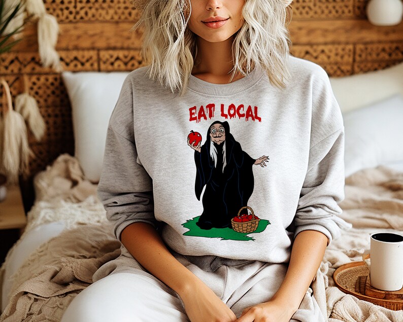 Puede incluir: Una sudadera gris claro con las palabras "EAT LOCAL" en rojo. El gr&aacute;fico presenta una bruja de dibujos animados sosteniendo una manzana roja y una cesta de manzanas. La bruja lleva una capa negra y est&aacute; de pie sobre hierba verde.