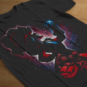 Puede incluir: Camiseta negra con un dise&ntilde;o de silueta de una figura sosteniendo una estrella. El dise&ntilde;o incorpora colores rojo, azul y blanco con un fondo tem&aacute;tico espacial y un barco rojo. La camiseta est&aacute; hecha de un material suave.