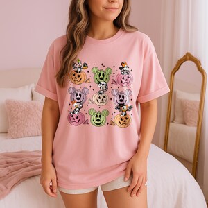 May include: A light pink t-shirt featuring a colorful Halloween-themed graphic. The graphic includes cartoon characters on pumpkins in various colors. The shirt has short sleeves and a relaxed fit, perfect for casual wear.