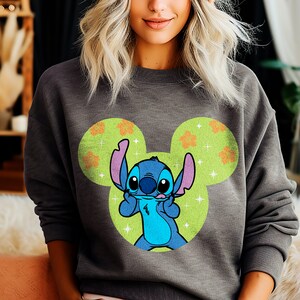 May include: Gray sweatshirt with a blue Stitch character wearing a Mickey Mouse ear hat. The hat is green with orange flowers and white sparkles.