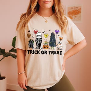 Puede incluir: Camiseta beige con un diseño de Halloween. El diseño presenta personajes de Star Wars como globos, incluyendo Darth Vader y un Stormtrooper, con el texto "TRICK OR TREAT". La camiseta es de estilo casual y cómodo.