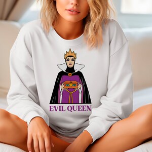 Puede incluir: Sudadera blanca con un gr&aacute;fico de la Reina Malvada de Blancanieves, sosteniendo un cofre del tesoro. La Reina Malvada est&aacute; representada con un vestido morado, capa negra y corona dorada. El texto "EVIL QUEEN" est&aacute; impreso debajo.