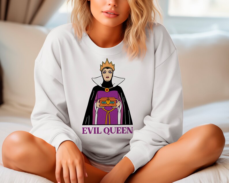 Puede incluir: Sudadera blanca con un gr&aacute;fico de la Reina Malvada de Blancanieves, sosteniendo un cofre del tesoro. La Reina Malvada est&aacute; representada con un vestido morado, capa negra y corona dorada. El texto "EVIL QUEEN" est&aacute; impreso debajo.