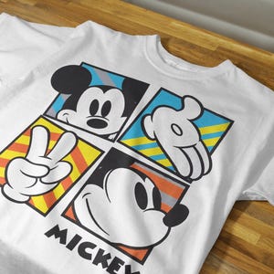 May include: White t-shirt with a colorful graphic design featuring Mickey Mouse in different poses. The design includes yellow, blue, red, and black colors. The text "MICKEY" is printed at the bottom of the shirt.
