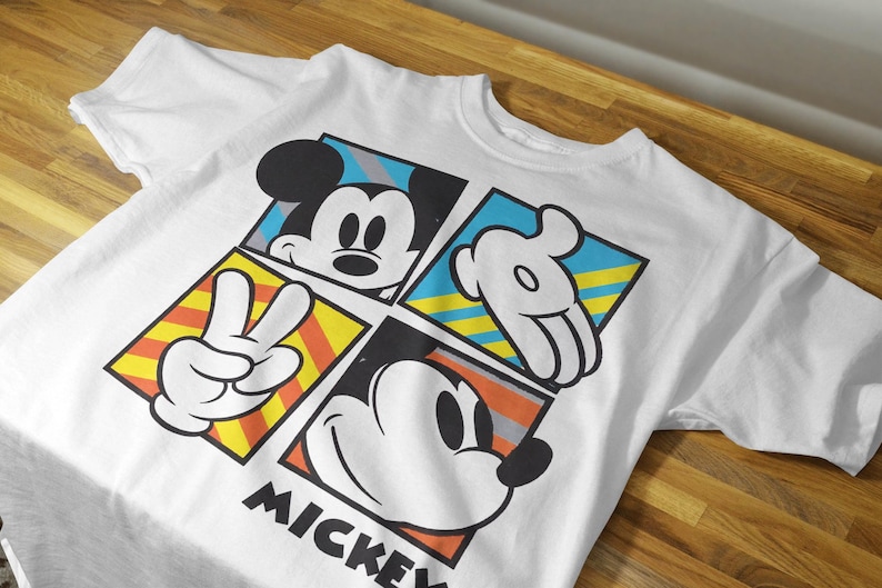 May include: White t-shirt with a colorful graphic design featuring Mickey Mouse in different poses. The design includes yellow, blue, red, and black colors. The text "MICKEY" is printed at the bottom of the shirt.