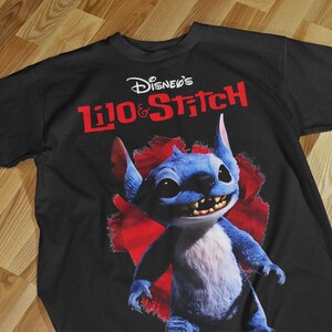 May include: Black t-shirt featuring the Disney's Lilo & Stitch logo in white and red. The shirt displays a vibrant image of Stitch, a blue alien creature, with a red background. The shirt is laid flat on a wooden surface.