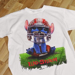 May include: White t-shirt featuring a blue cartoon character wearing a football helmet, sitting on a football. The graphic includes the text "Lilo & Stitch" in red. The shirt is on a wooden surface.