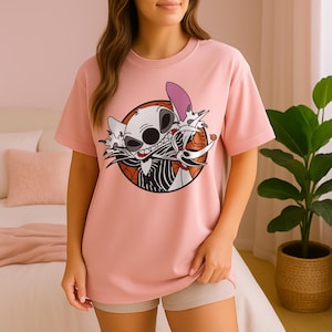 Può includere: T-shirt rosa chiaro con una grafica di un personaggio dei cartoni animati con un volto a forma di teschio, un orecchio viola e un abito a righe bianche e nere. La grafica è all'interno di un cerchio arancione e marrone.