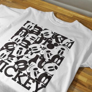 May include: White t-shirt with a black and white graphic print of Mickey Mouse heads arranged in a square pattern. The text "MICKEY" is printed in black below the graphic.