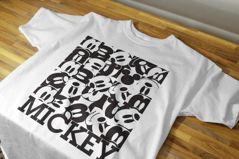 May include: White t-shirt with a black and white graphic print of Mickey Mouse heads arranged in a square pattern. The text "MICKEY" is printed in black below the graphic.