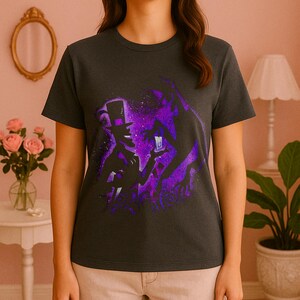 May include: Dark gray t-shirt featuring a purple and black graphic of a magician in a top hat holding a glowing object. The design has a starry background and a silhouette of a figure. The shirt is a crew neck and has short sleeves.