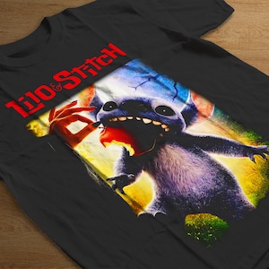 Può includere: T-shirt nera con grafica di Stitch di Lilo & Stitch. Il disegno mostra Stitch che mangia una mela, con le parole "Lilo & Stitch" in rosso. L'immagine ha uno stile colorato e a cartoni animati.