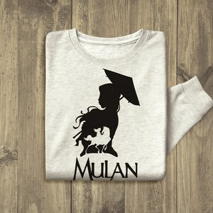 Op de afbeelding: Een grijs longsleeve t-shirt met een zwart silhouet van een vrouw die een paraplu en een draak vasthoudt. De tekst "MULAN" is onder het silhouet gedrukt.