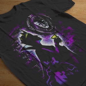 Puede incluir: Camiseta negra con una silueta de un personaje sosteniendo una copa, con un espejo y una cara en el fondo. El dise&ntilde;o incorpora acentos morados y blancos, con un tema c&oacute;smico y un toque de fantas&iacute;a.