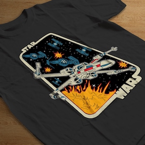 Puede incluir: Camiseta negra con un diseño de Star Wars. El gráfico incluye un caza X-wing, otras naves espaciales y las palabras "STAR WARS" en un estilo retro. El diseño está ambientado sobre un fondo estrellado con llamas.
