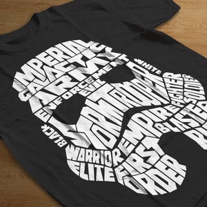 Puede incluir: Camiseta negra con un diseño blanco de casco de Stormtrooper compuesto por palabras como "Imperial", "Empire" y "Warrior". El diseño es una ilustración tipográfica única.