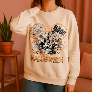 Puede incluir: Sudadera color crema con un diseño de Halloween. El diseño incluye a Mickey Mouse, Minnie Mouse, Donald Duck y Goofy con disfraces de Halloween. La palabra "HALLOWEEN" está impresa en naranja debajo de los personajes.