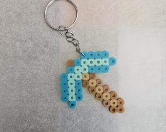 May include: A pixelated pickaxe keychain made of blue and tan plastic beads. The pickaxe is attached to a silver-colored keyring and chain. The keychain is a handmade item.