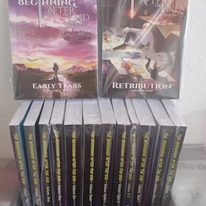 Puede incluir: Una colección de libros "The Beginning After The End". Dos volúmenes se muestran en posición vertical, con "Early Years" y "Retribution" visibles. Los otros volúmenes están apilados debajo, con el título de la serie y los números de volumen claramente impresos.
