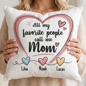 Puede incluir: Cojín decorativo blanco con un diseño de corazón rosa y el texto "All my favorite people call me Mom." Debajo del corazón hay tres corazones más pequeños en azul, rosa y amarillo, cada uno con un nombre: Liha, Noah y Lucas.
