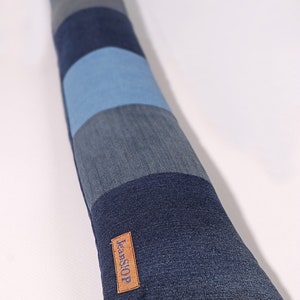 Op de afbeelding: Een lange, rechthoekige deurstop van denim met een bruin leren label waarop "JeanSTOP" staat. De deurstop is gemaakt van verschillende tinten blauwe denim.