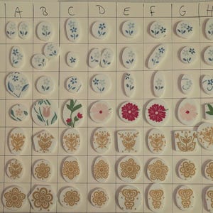 May include: A grid of small, white, decorative ceramic pieces with various floral and geometric designs. The pieces feature blue, pink, and gold accents. Shapes include ovals, circles, and rectangles. Some have a single flower, others clusters.