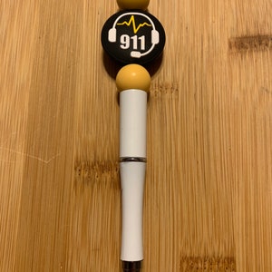 May include: White pen with a black and white 911 emergency services logo charm with a yellow heart beat graphic. The charm is surrounded by three yellow beads.