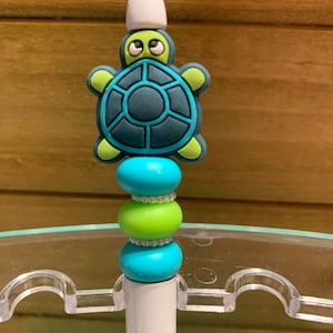 May include: A white pen with a green and blue turtle topper and three blue and green beads. The pen is in a clear plastic holder.