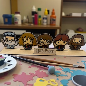 May include: A wooden Harry Potter-themed display featuring five cartoon figures: Dumbledore, Hermione, Harry, Ron, and Hagrid. The figures stand on a wooden base with the words "Harry Potter" and are painted in various colors. Two paintbrushes and a plate are in the foreground.