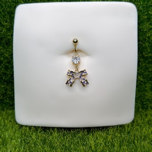May include: A gold-colored belly button ring with a clear gem at the top and a bow-shaped charm. The bow charm has purple and clear accents. The ring is displayed on a white surface against a green background.