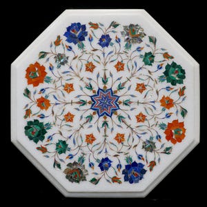 Puede incluir: Mesa octogonal de mármol blanco con diseño floral incrustado. El intrincado patrón presenta flores y estrellas de color naranja, azul, verde y plateado. La mesa es una pieza decorativa.