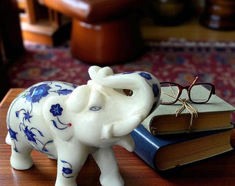May include: A white elephant figurine with blue floral designs. The elephant has its trunk raised and is standing on a wooden surface. A pair of eyeglasses rests on a stack of books. The figurine is a decorative item.